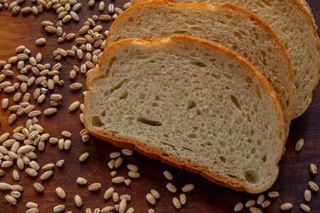 Slices of sliced white bread on a dark board and spilled grains of wheat