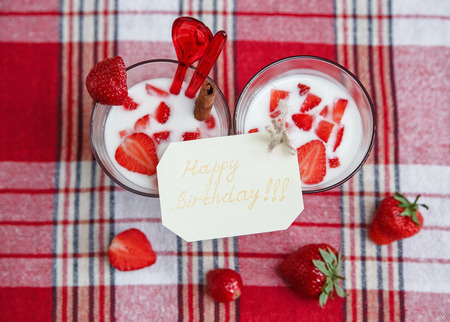 Two Glasses of Milk Cocktail,Red Fresh Strawberries with Plastic Spoons and Cinnamon on the Check Tablecloth.Wish Birthday  Card.Breakfast Organic Healthy Tasty Food.Cooking Vitamins Ingredients.Summer Fruits.Selective Focusの写真素材
