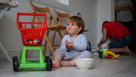 Cute little baby girl sitting on hardwood near play stroller and smiling. Child keep palms together...