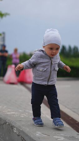 Baby boy in white knitted hat walking on parapet in the squareの写真素材