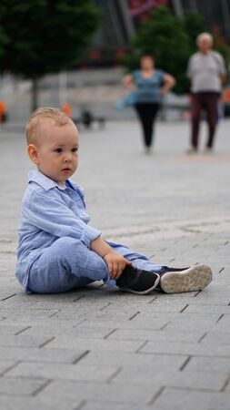 Little funny boy in blue clothes sitting on the pavement slabs on the squareの写真素材