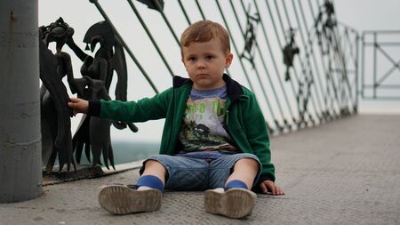 Little funny boy in green jacket sitting on the metal bridge in the park.の写真素材