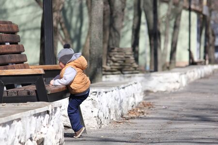 Cute child in orange jacket climbing on the bench in the spring park