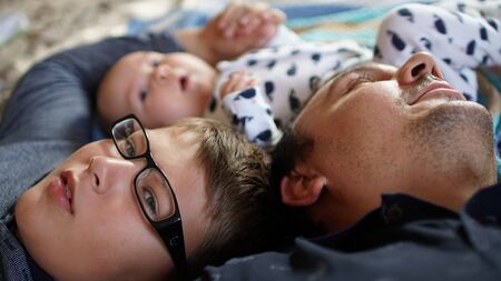 Portrait of young man with his sons lying on the bed at home . Close up of happy father, his cute toddler and son-teenager with glasses .の写真素材