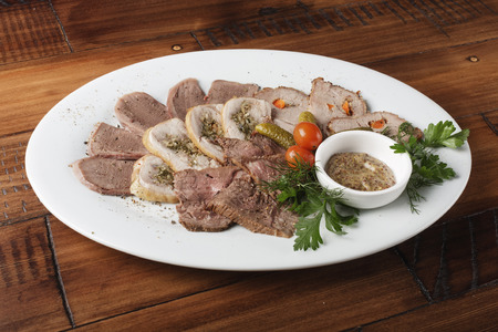 Baked roast beef, chicken rolls, tongue and pork meat with vegetables in a white oval plate.の写真素材