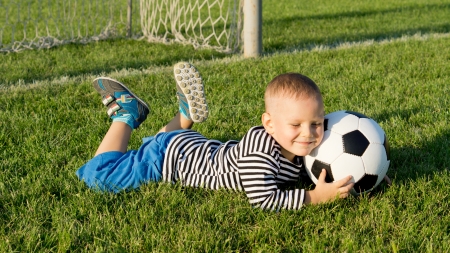 Smiling happy young boy lying on green grass at a sportsfield in evening light with a soccer ballの写真素材