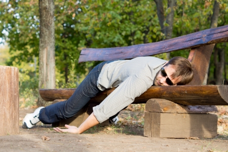 Exhausted man in casual clothing and sunglasses sleeping on a rustic wooden bench in woodlandの写真素材