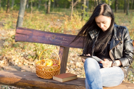 Young casual woman sitting on a rustic wooden park bench with a basket of apples and book using a tabletの写真素材