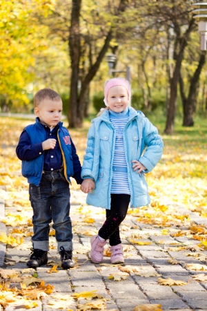 Young brother and sister holding hands and standing posing on a paved walkway in an autumn park with colouful yellow leavesの写真素材