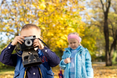 Little boy playing with a vintage slr camera in a colourful autumn park watched by his little sisterの写真素材