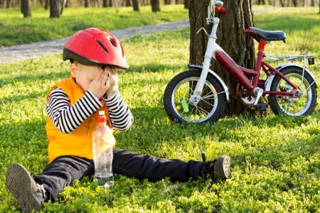 Embarassed little boy in a safety helmet hiding his face from the photographer while sitting on lush green grass drinking bottled water with his bicycle propped up against a tree alongside himの写真素材