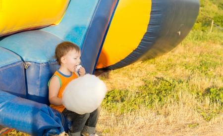 Young blond boy eating cotton-candy and other candies sitting near a colorful inflatable-slideの写真素材