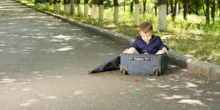 Little boy playing in a suitcase lying at the side of a tarred country road lined with leafy green treesの写真素材