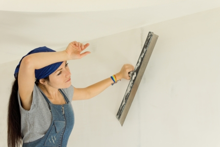 Tired woman wallpapering a wall pausing for a breather to wipe her forehead with her hand when doing home renovationsの写真素材