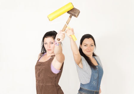 Two young women in overalls posing with a paint roller and wooden mallet in their raised hands smiling at the cameraの写真素材