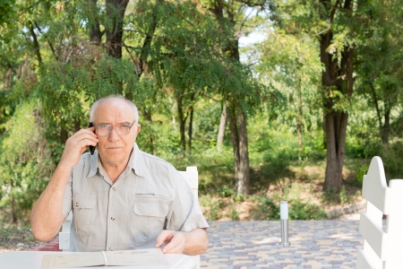 Elderly man chatting on his mobile phone sitting outdoors at a restaurant table in the shade of a treeの写真素材