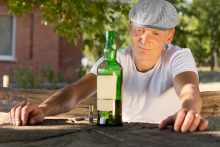 Melancholic drunk Caucasian man sitting at a table in the park and looking at a bottle of wineの写真素材