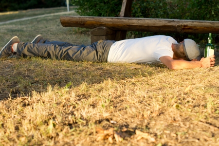 Drunk man sleeping on the ground next to a bench with a hand on a bottle of white wineの写真素材