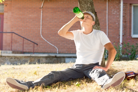 Alcoholic man sitting drinking under a tree gulping down spirits straight from the bottleの写真素材