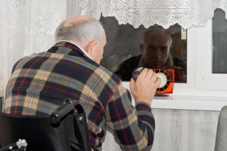 Handicapped man in a wheelchair sitting with a clock in front of him waiting patiently at a window at night for someone to arriveの写真素材