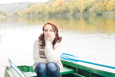 Pretty young woman sitting in a rowboat on a tranquil autumn lake with her chin on her hand giving the camera a thoughtful lookの写真素材
