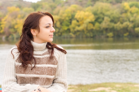 Beautiful girl enjoying the autumn weather sitting outdoors with a lovely smile on her face in a warm polo-neck sweater in front of a tranquil lake or riverの写真素材