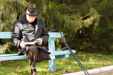 Elderly handicapped man on crutches with only one leg having suffered an amputation sitting in the sun reading his book on a park bench in a warm coat and hatの写真素材
