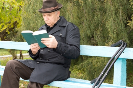 Disabled man on crutches sitting reading in the sun on a rustic wooden bench in the park in his warm overcoat and hat as he enjoys the last sunshine of the autumnの写真素材