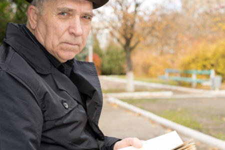 Close up portrait of an elderly man in an overcoat and hat sitting outdoors in the autumn sunshine in a park enjoying the peace while reading his book looking at the camera with a serious expressionの写真素材
