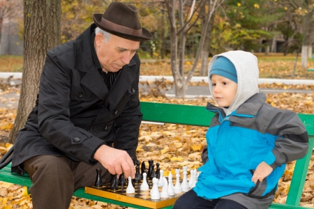 Little boy playing chess with his grandfather sitting back comfortably on a park bench waiting for his grandfather to make his moveの写真素材