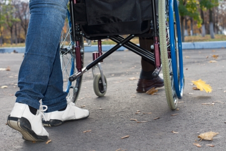 Close up of the legs of a carer in jeans and sneakers pushing a wheelchair along a tarred road, low angle viewの写真素材