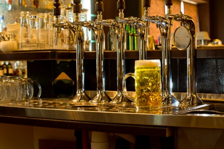 Row of stainless steel beer taps on a wooden counter for dispensing draught beer in a pub with a large glass tankard of beer alongsideの写真素材