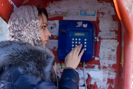 Beautiful young woman standing dialling a number to make a connection on a public telephone in a booth alongside an urban streetの写真素材
