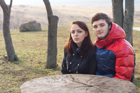 Attractive young couple sitting together at a rustic picnic table in a public park in the country wrapped up warmly against the cold misty winter weatherの写真素材