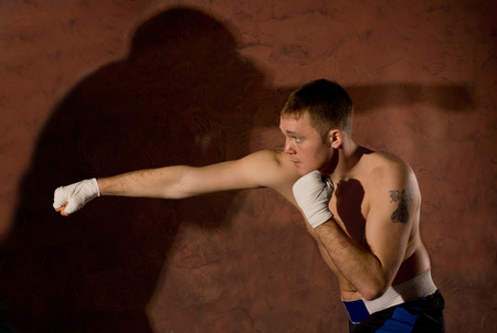 Young boxer punching an opponent during a fight in the ring with his fist extended and the shadow of his opponent cast on the wall behind himの写真素材