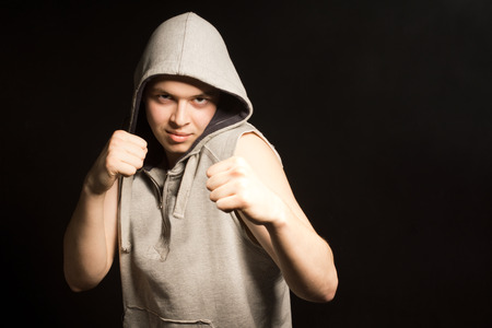 Determined angry young boxer in a hooded top raising his bare fists in a threatening gesture on a dark with copyspaceの写真素材