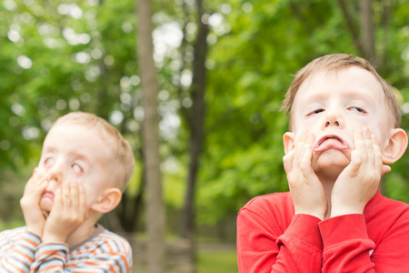 Two young boys playing outdoors together in a park or garden standing pulling funny faces using...