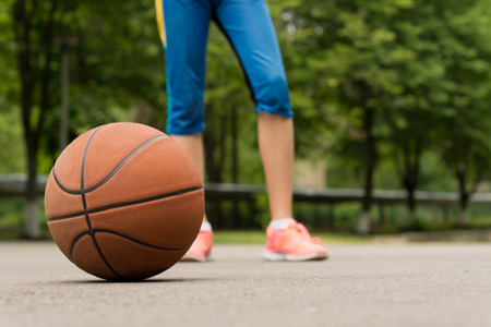 Basketball on an outdoor asphalt court with the legs of an athletic slender young girl player behind against a backdrop of leafy green treesの写真素材
