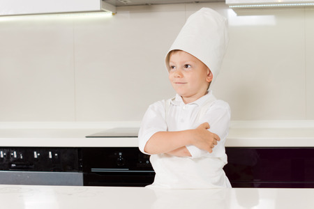 Confident smiling young boy in a chefs uniform standing in the kitchen in front of the stove with his arms foldedの写真素材