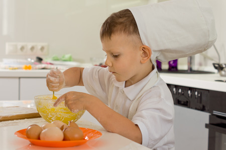 Cute young boy chef in a white toque standing at a kitchen counter whipping the eggs in the mixing bowlの写真素材