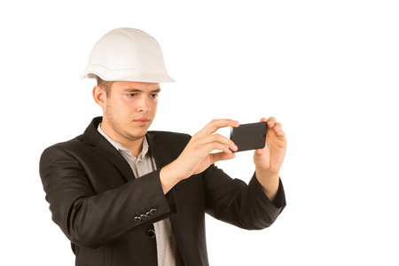 Close up Young Male Engineer in White Helmet and Black Coat Taking Picture Using Mobile Phone. Isolated on White Background.の写真素材