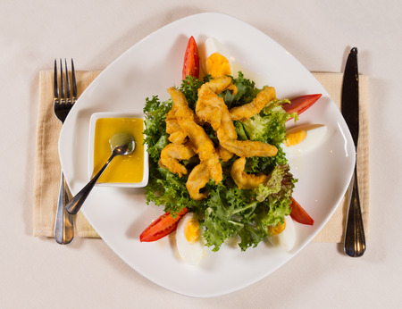 Close up Gourmet Tasty Fried Chicken Meat Dish on Lettuce with Egg, Tomatoes and Dipping Sauce. Served on White Plate with Utensils on Sides for Guest.の写真素材