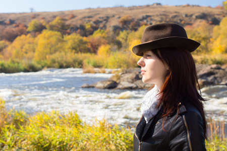 Attractive stylish woman with long brown hair wearing a trendy hat, scarf and jacket posing in countryside in front of a river, with copyspace, side viewの写真素材