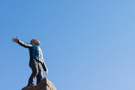 Exuberant young man on top of an old stone wall standing with his arms outspread shouting at the sun against a clear blue sky with copyspace, view looking up from belowの写真素材