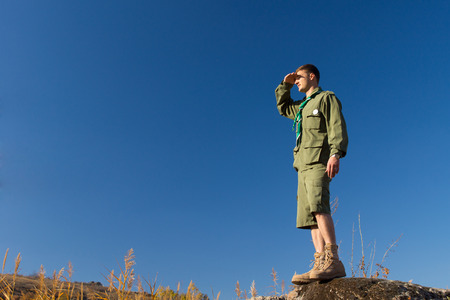 Young Male Scout Standing on Big Rock Observing the Field at the Camp Area on a Blue Sky Background.