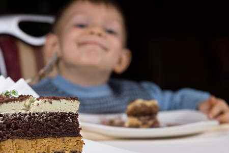 Close up of a slice of freshly baked layer cake for dessert on a table with a playful little boy grinning with glee behind itの写真素材