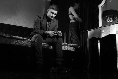 Wedding proposal rejected concept with a young man sitting on the end of a bed despondently looking at the engagement ring as his girlfriend paces around in the backgroundの写真素材