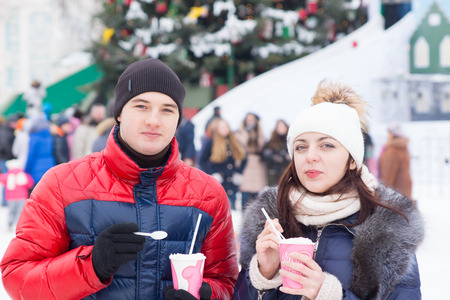 Young couple enjoying a Christmas carnival on a cold snowy winter day standing outdoors drinking takeaway coffee with a crowd of people and decorated pine tree in the backgroundの写真素材