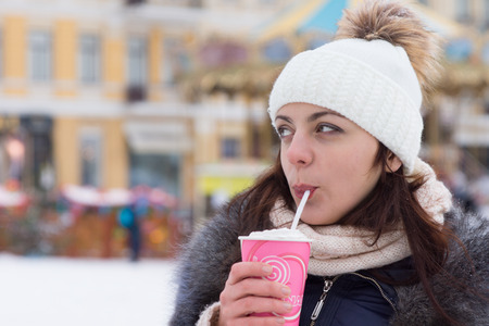 Young woman sipping a hot drink in winter weather standing outdoors in a snowy town square in her knitted hat and scarf looking to the side watching somethingの写真素材