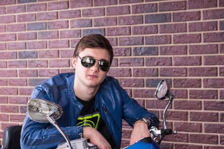 Trendy young man on a motorbike leaning over the handlebars in his fashionable sunglasses to smile...